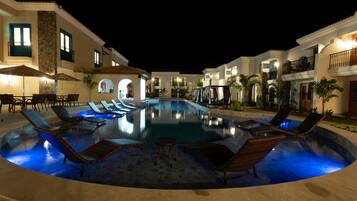 Un área de piscina nocturna con sillones de descanso y sombrillas, rodeada de edificios con balcones e iluminación exterior.