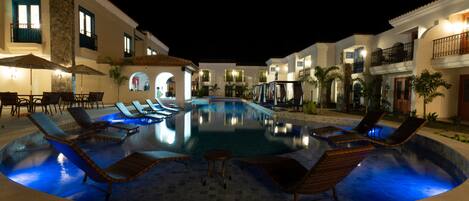 A nighttime swimming pool area with lounge chairs and umbrellas, surrounded by buildings with balconies and outdoor lighting.