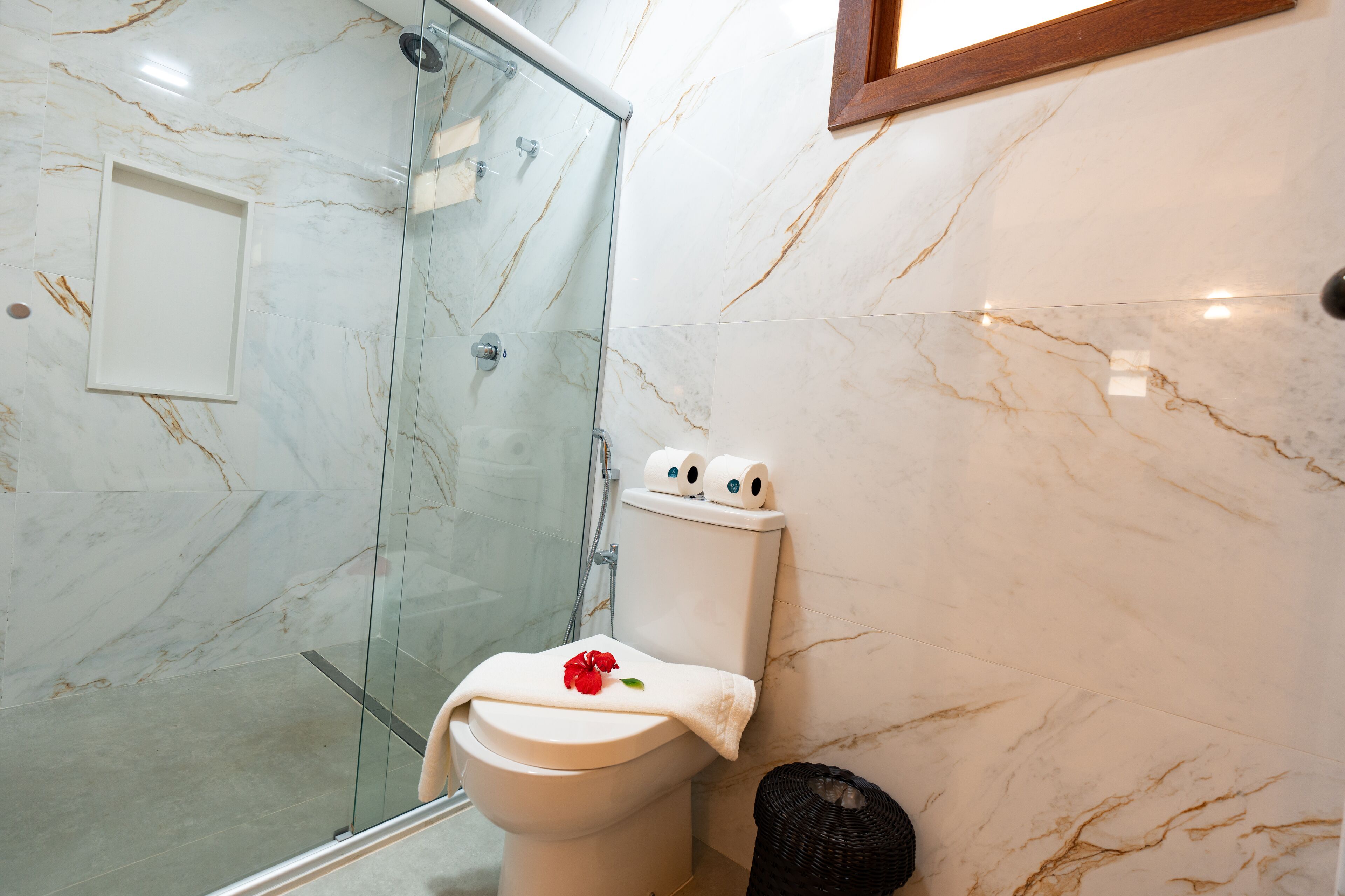A bathroom with a glass shower enclosure, a toilet with a decorative towel, and a trash bin.