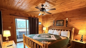 A wooden cabin bedroom with a bed, two bedside lamps, a ceiling fan, and a window with curtains.