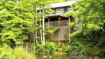 Eine Holzhütte mit einer Holzterrasse, umgeben von dichtem Grün und einem Teich mit Enten.
