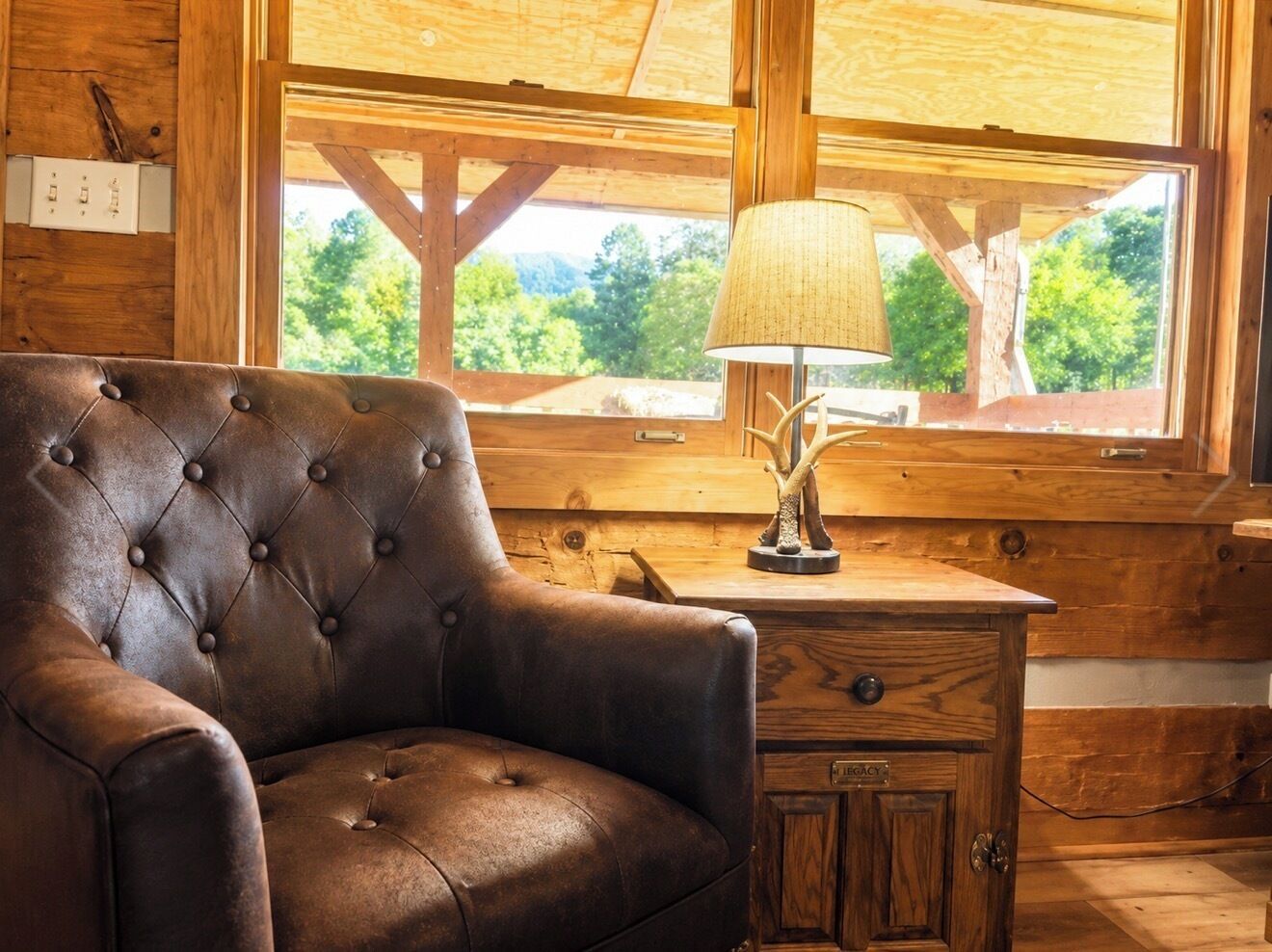 A wooden cabin interior with a leather armchair, a lamp, and a window with a view of trees.