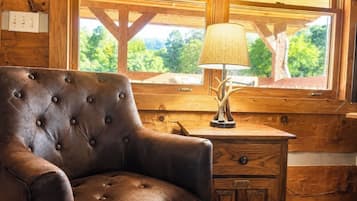 A wooden cabin interior with a leather armchair, a lamp, and a window with a view of trees.