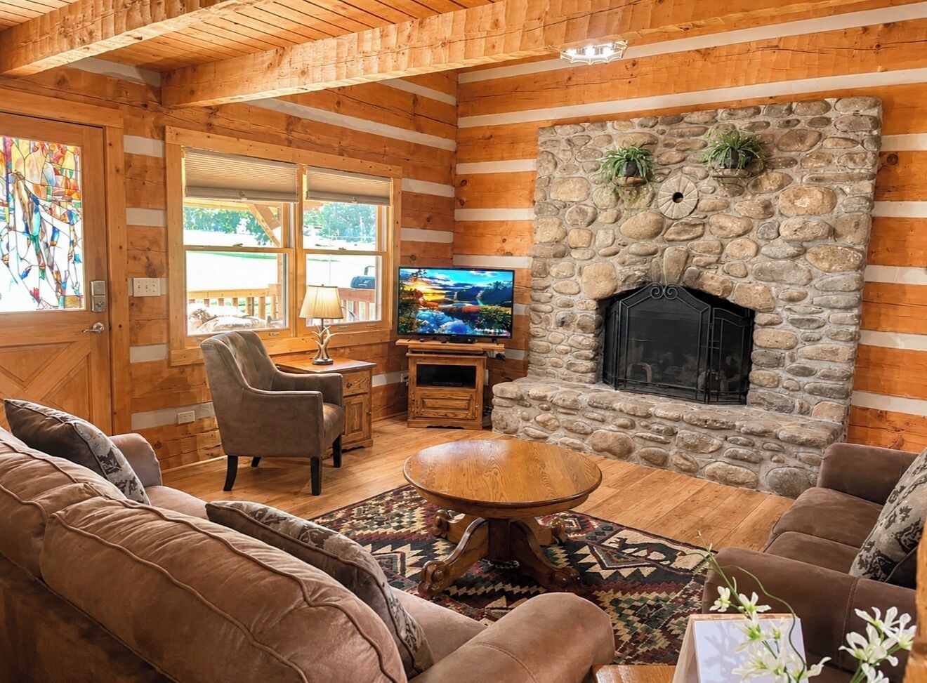 A cozy log cabin interior with a stone fireplace, wooden furniture, and a television.