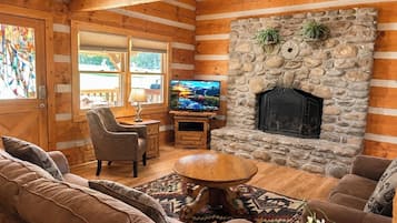 A cozy log cabin interior with a stone fireplace, wooden furniture, and a television.