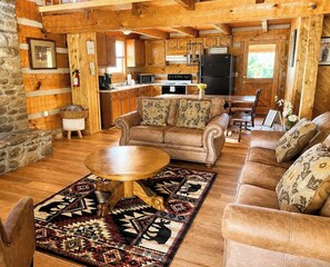 A log cabin interior with a living room, sofa, coffee table, rug, fireplace, kitchen area, and stone wall.