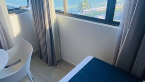 Ett hotellrum med en säng, en stol, ett stort fönster med utsikt över havet och en palm.