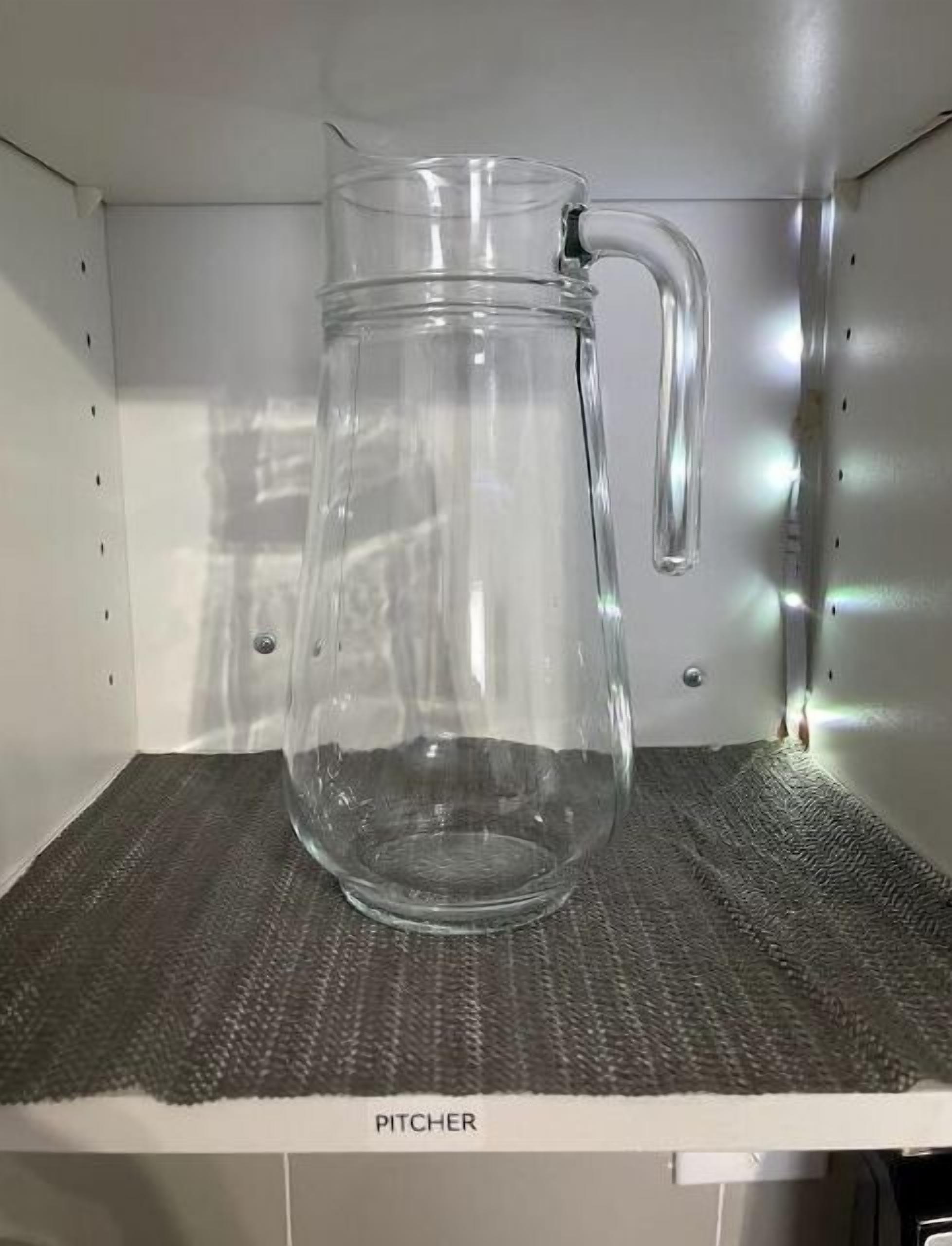 A clear glass pitcher on a shelf with a textured mat labeled "PITCHER".