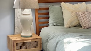 Ein Bett mit hölzernem Kopfteil, ein Nachttisch mit Lampe und ein Fenster mit Vorhängen.