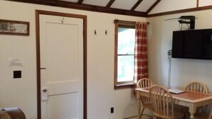 A cozy room with a checkered floor, a wooden table with chairs, a television mounted on the wall, and a door with a small window.