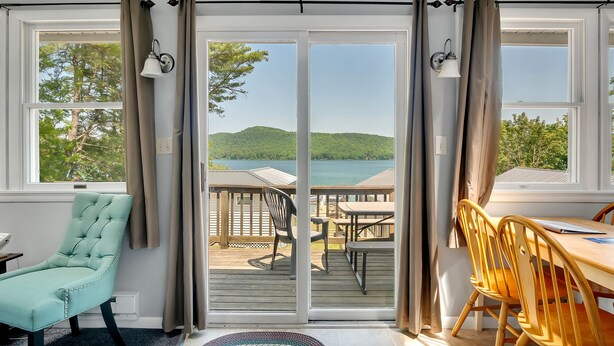 A room with a view of a lake and hills, a sliding glass door, a teal chair, a wooden table, and a yellow chair. - 3 Bedroom Cottage only 10 minutes from Cooperstown (Cooperstown)