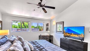Une chambre à coucher comprenant un lit, un bureau, une chaise, un téléviseur et des fenêtres donnant sur un paysage verdoyant.