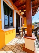 Um espaço ao ar livre coberto, com chão de azulejos, uma mesa de madeira e cadeiras.