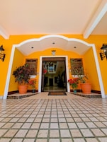 Entrada de hotel com piso de azulejos, arco central e plantas em vasos de ambos os lados.