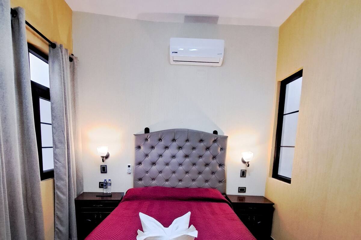 Habitación de hotel con cabecera capitonada, colcha roja, toalla blanca doblada con cuidado, dos lámparas de noche y un equipo de aire acondicionado en la pared.