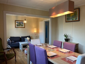 Un coin repas avec une table en bois dressée pour quatre personnes, entourée de chaises violettes. On aperçoit un canapé et une petite table avec une lampe à l’arrière-plan.
