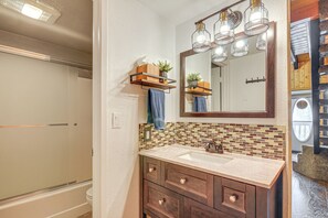 Une salle de bain dotée d’une douche à parois de verre, d’un lavabo beige avec un meuble-vasque en bois, d’un miroir avec des appliques murales et d’un pare-feu en carreaux.