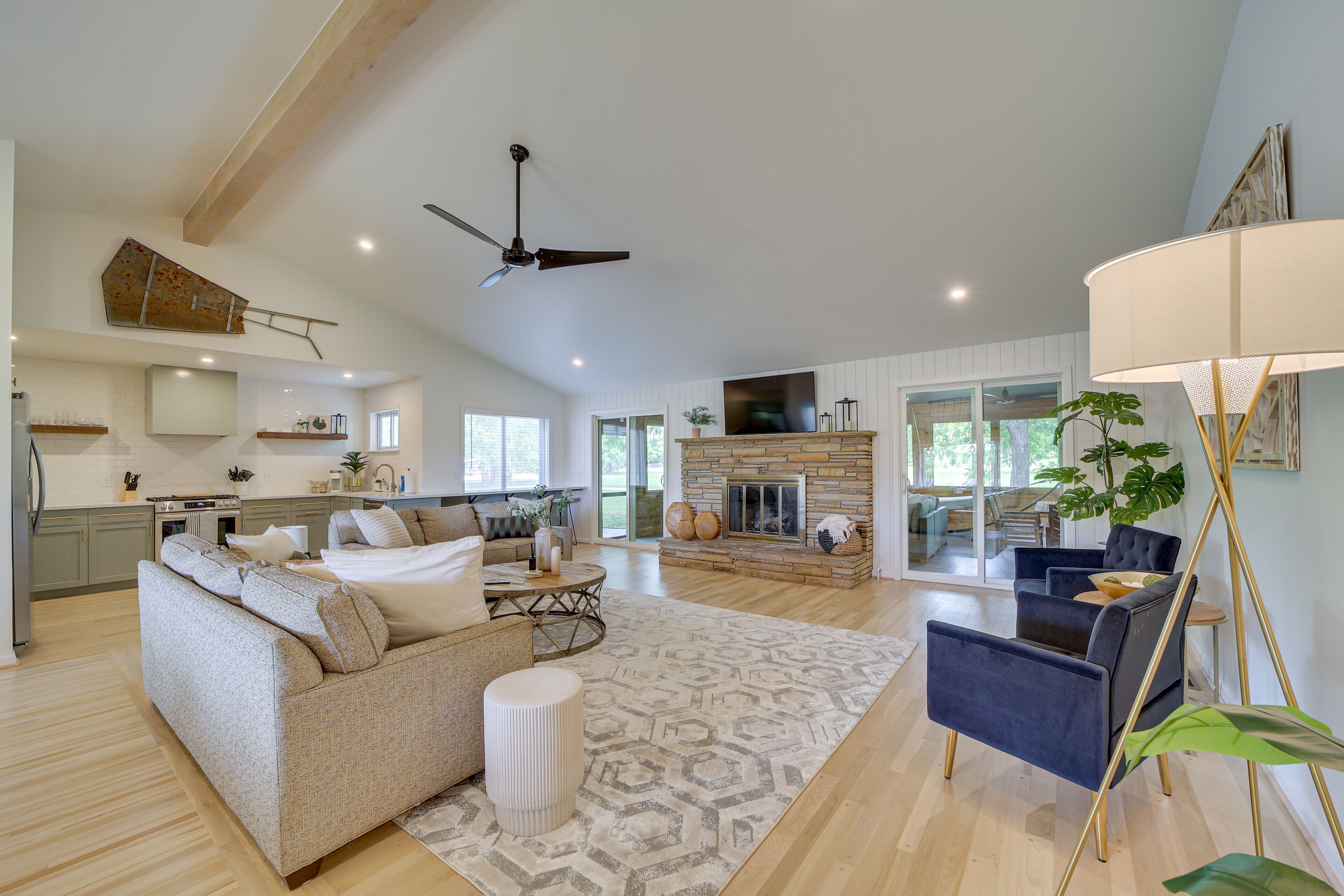 A spacious living area with a sofa, armchair, and a fireplace. There is a ceiling fan and a kitchen in the background.