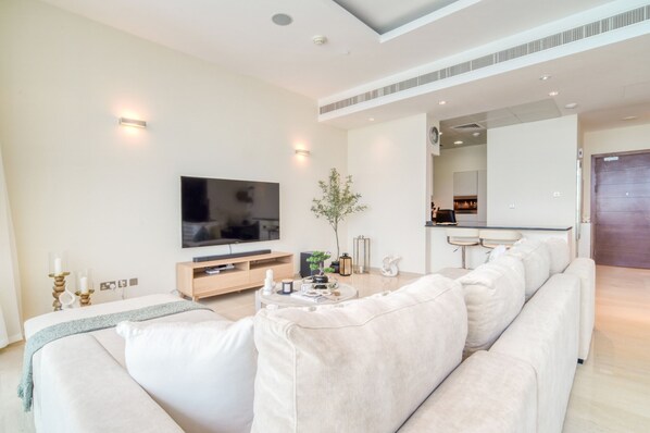 A modern living room with a large flat-screen TV, a sofa, a coffee table, and a small kitchen area in the background.