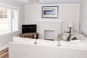 A living room with a white brick fireplace, a flat-screen TV on a wooden stand, a white sofa with cushions, and a framed picture above the fireplace.
