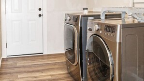 A laundry room with two front-loading washing machines and a dryer, wooden flooring, and a white door. - Salem Road Ranch | 3 BR w/ Outdor Firepit and Backyard (Lincolnton)