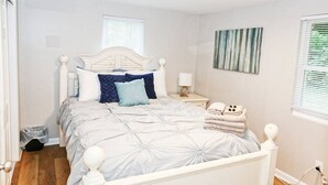 Un lit bien fait, avec une tête de lit blanche, une table de chevet avec une lampe et une fenêtre équipée de stores.