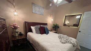 A neatly arranged bedroom with a large bed, a bedside table with a lamp, a dresser, a mirror, and a potted plant.