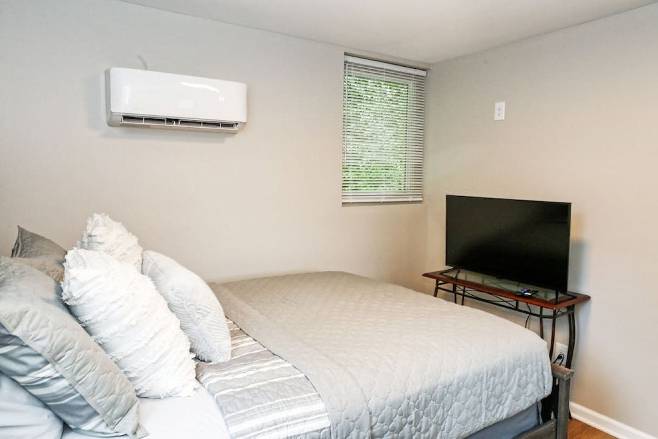 Un dormitorio con una cama, un televisor en un soporte, un equipo de aire acondicionado y una ventana con persianas.