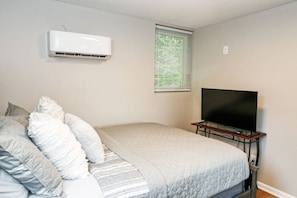 Une chambre à coucher avec un lit, un téléviseur posé sur un meuble, un appareil de climatisation et une fenêtre avec des stores.