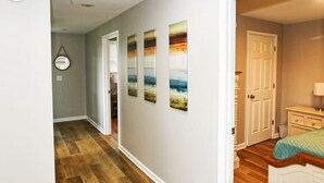 Un couloir doté d’un plancher en bois, une porte donnant sur une chambre, et trois tableaux représentant des paysages accrochés au mur.