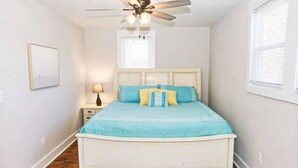 A bedroom with a white bed, a ceiling fan, a nightstand with a lamp, and a window with blinds.