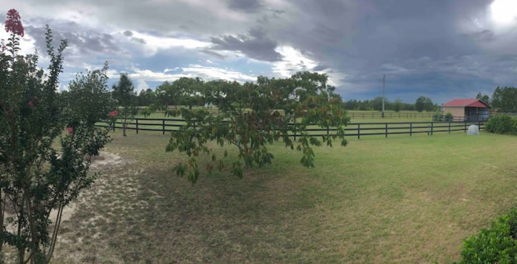 A rural landscape with a fenced pasture, a red-roofed building, and a cloudy sky.
