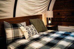Una cama con manta escocesa y varias almohadas, cabecera de madera, mesita de noche con lámpara y cómoda blanca.