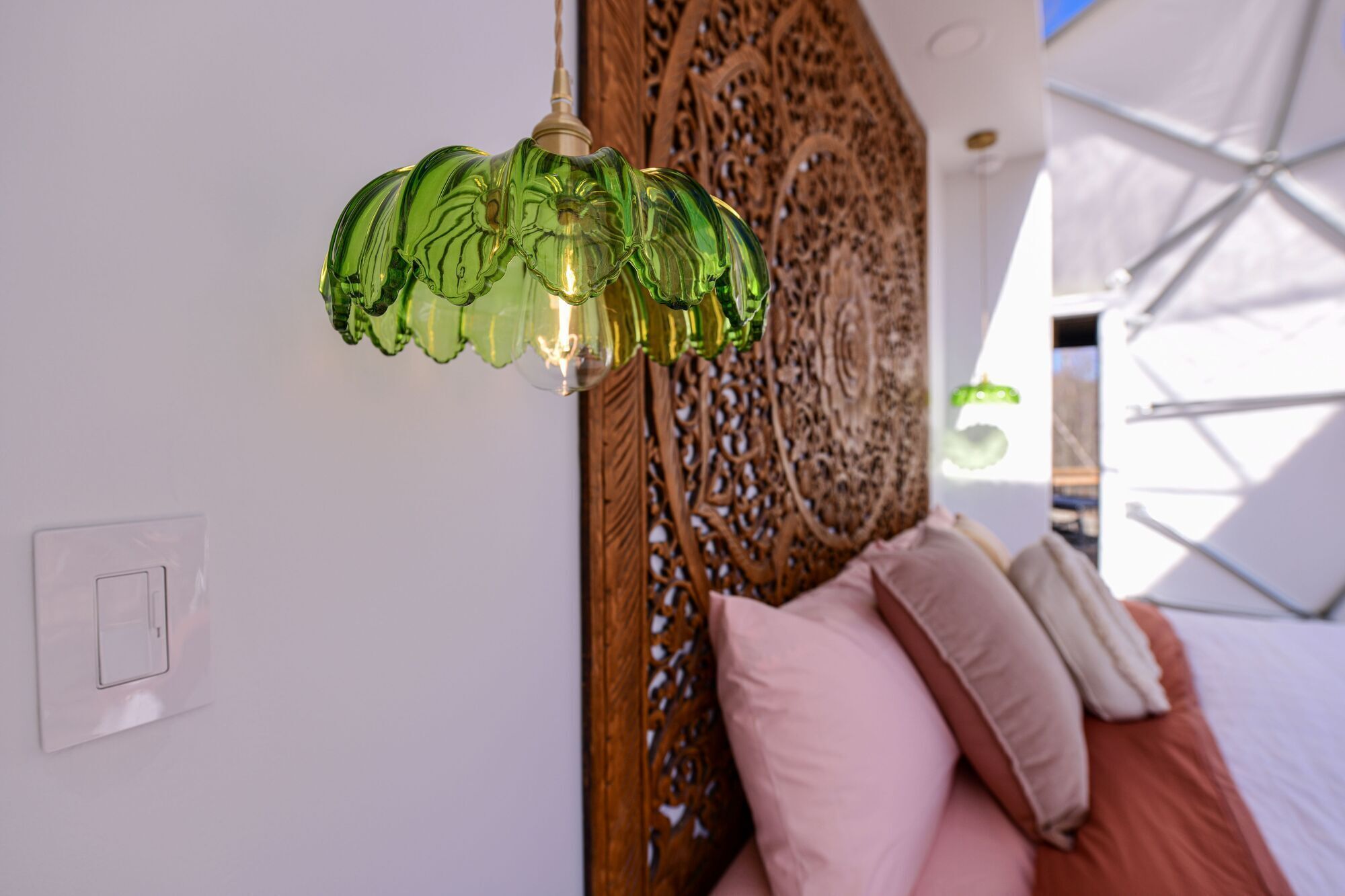 A room with a green glass lamp, a wooden carved headboard, and a bed with pink and beige pillows.