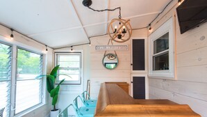 Une cuisine avec des armoires en bois, un plan de travail en bois et un luminaire suspendu. On y trouve une fenêtre avec des stores, une plante en pot et un panneau accroché au mur.