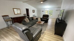Amplia sala de estar con un sofá de cuero, una mesa de centro de madera y un sillón de cuero negro. Se observa un ventilador de techo, un mueble para televisor y una puerta que conduce a otra habitación.