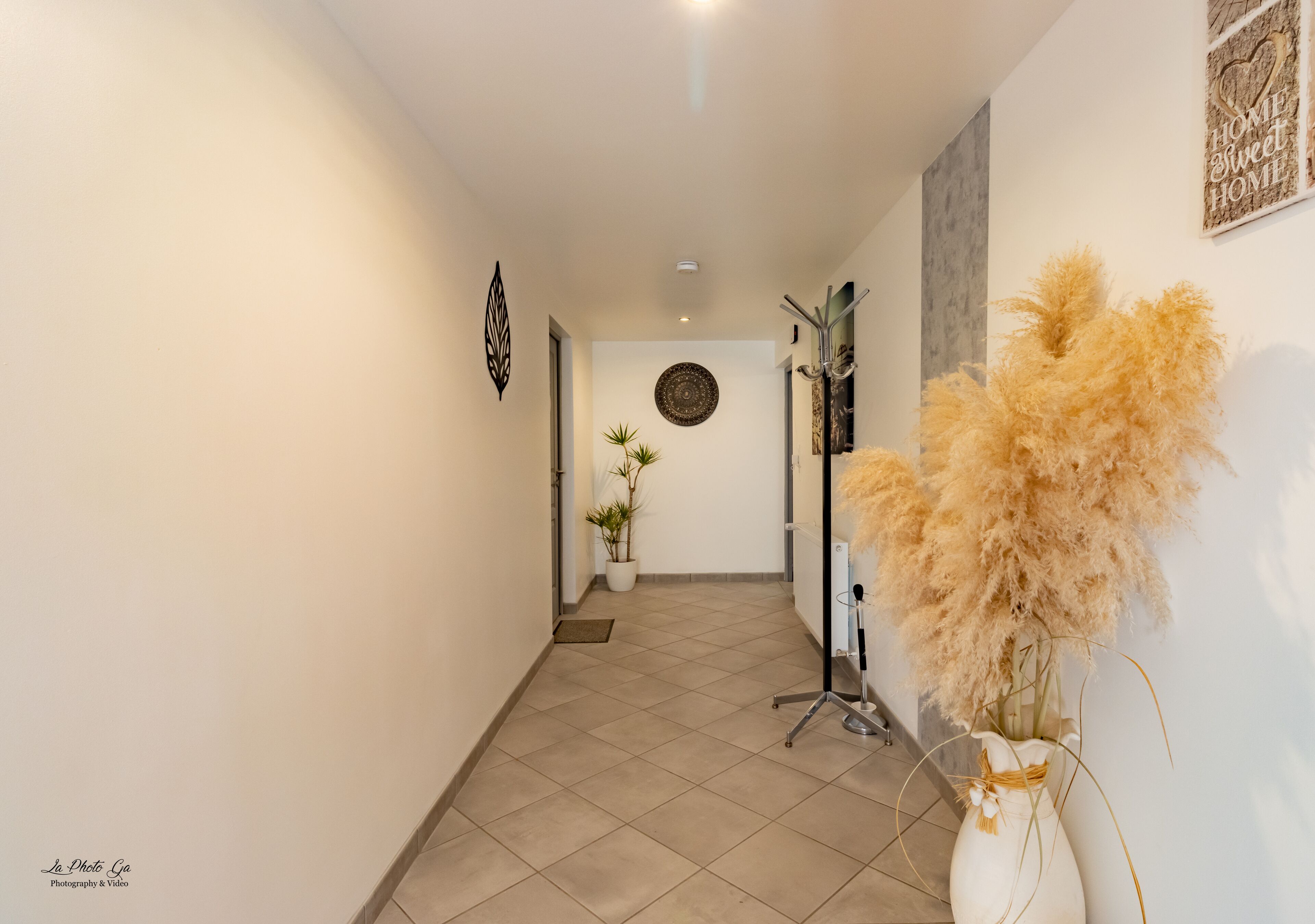 Un couloir doté d’un sol carrelé, d’une plante en pot, d’un élément décoratif mural et d’un grand vase contenant des plumes d’herbe des pampas.
