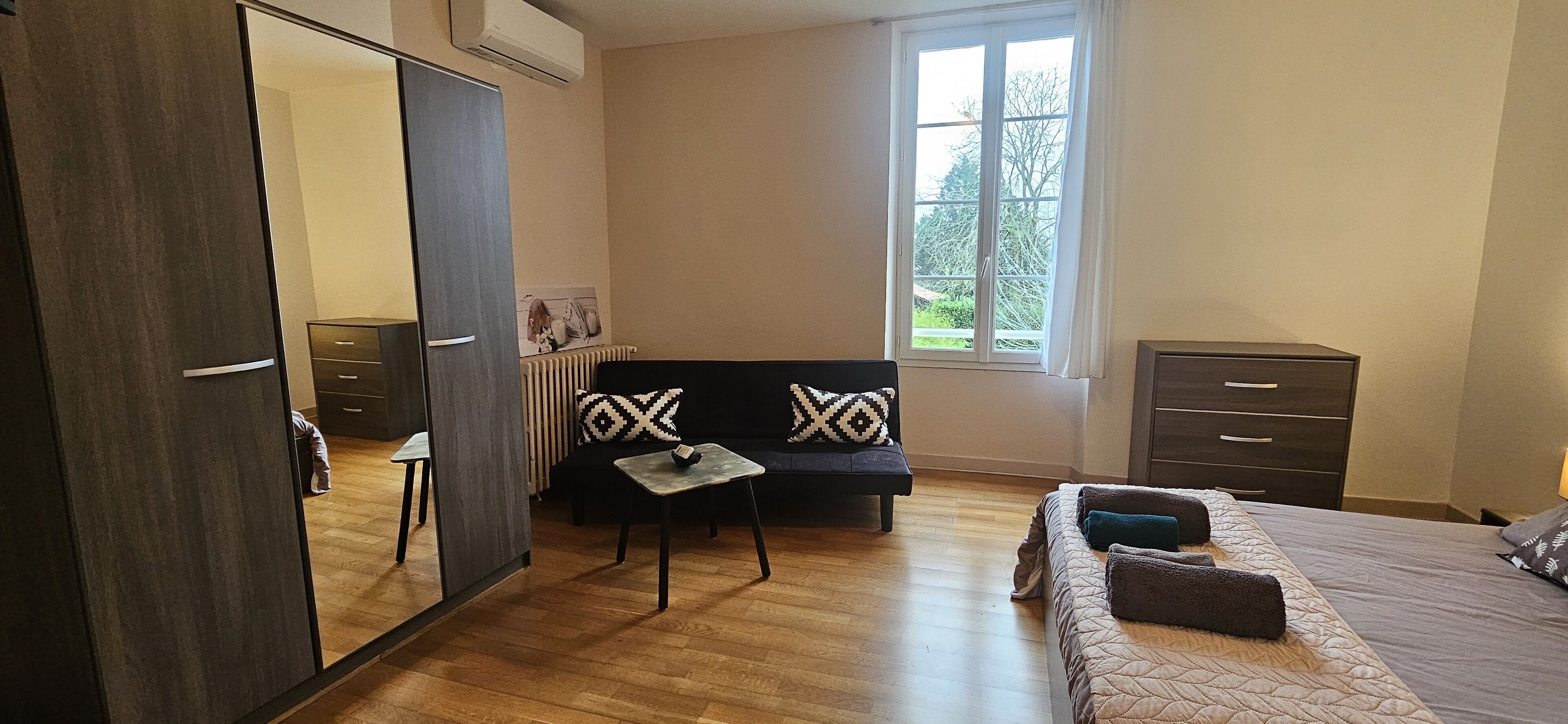 A bedroom with a bed, a sofa, a small table, a wardrobe, a radiator, and a window with a view of greenery.