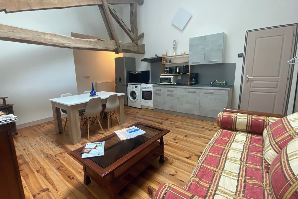 A cozy living area with a wooden floor, a sofa, a coffee table, and a dining table with chairs. A kitchen with appliances is visible in the background.