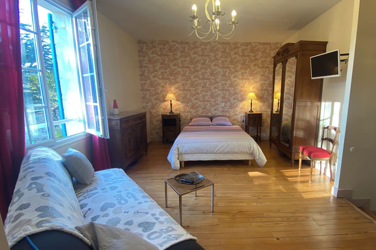 A bedroom with a large bed, a wooden floor, a sofa, a TV, and a window with curtains.