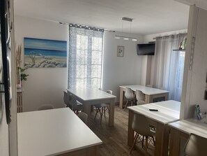 Een moderne eethoek met witte tafels en stoelen, een schilderij van een strandgezicht, een aan de muur bevestigde televisie en een raam met dunne gordijnen.