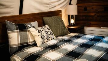 Una cama con manta escocesa y varias almohadas, cabecera de madera, mesita de noche con lámpara y cómoda blanca.