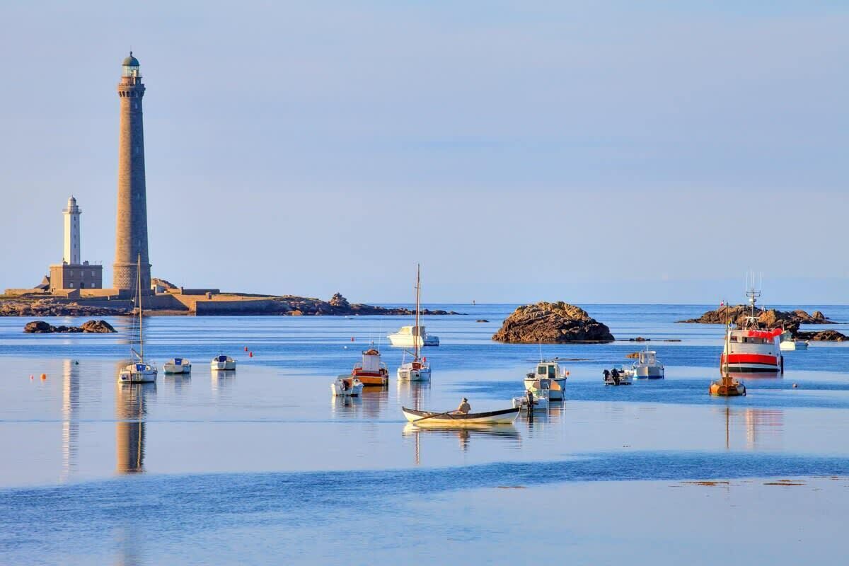 A coastal scene with a lighthouse, calm water, and several boats.