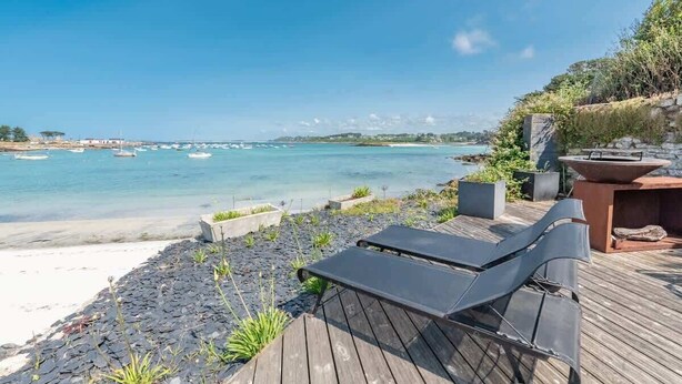 Una terraza de madera con sillones de descanso con vistas a una playa donde se ven barcos en el agua.