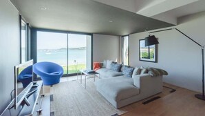Una sala moderna con un ventanal grande que ofrece vista al mar, un sofá gris, un sillón azul y un escritorio con una computadora.
