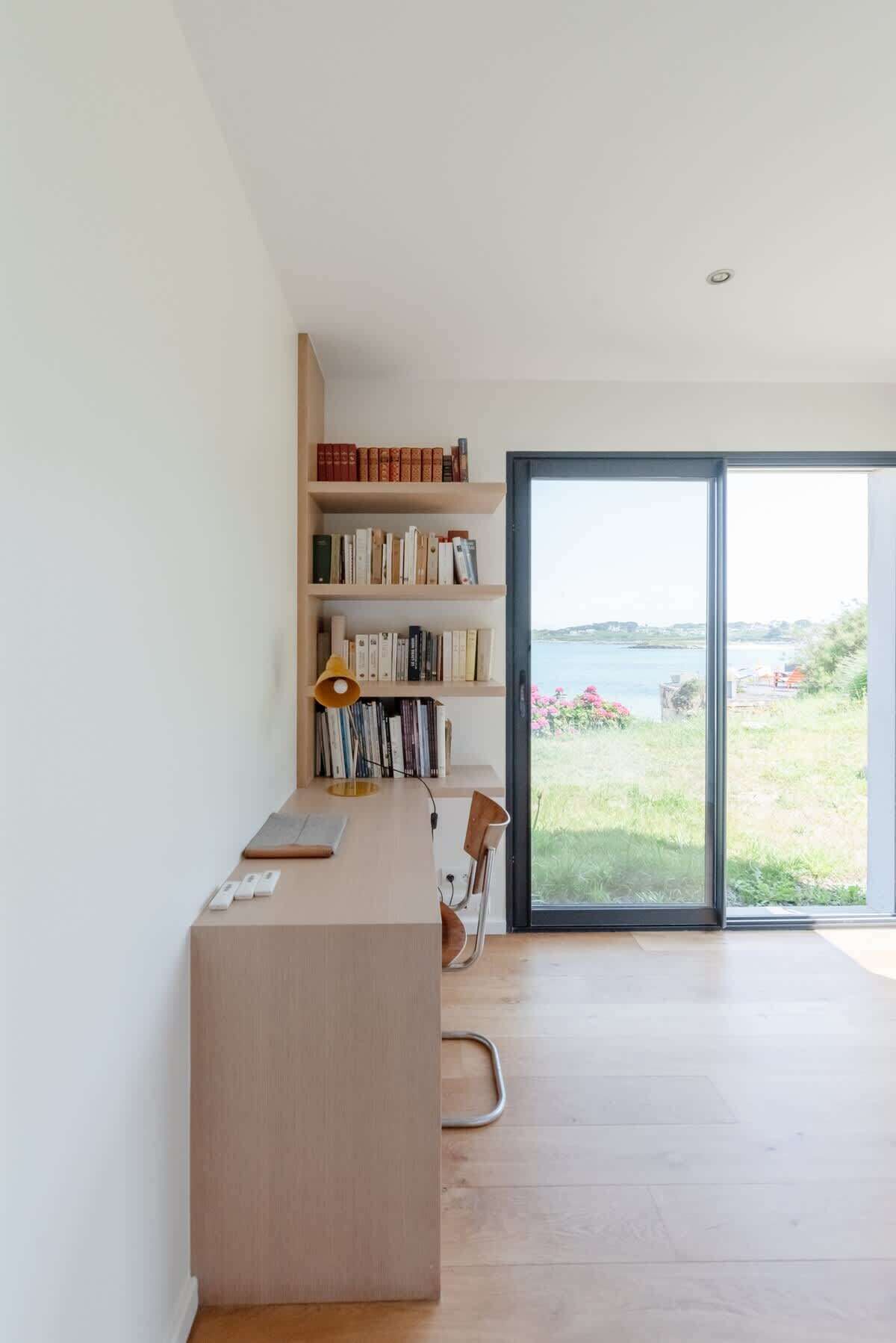 A modern, minimalist room with a wooden desk, bookshelves, and a large sliding glass door leading to a scenic outdoor view.