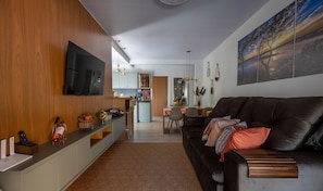 A modern living room with a flat-screen TV, a dark leather sofa, a dining area, and a kitchen in the background. - Ibira Oásis - Imbituba - SC (Imbituba)