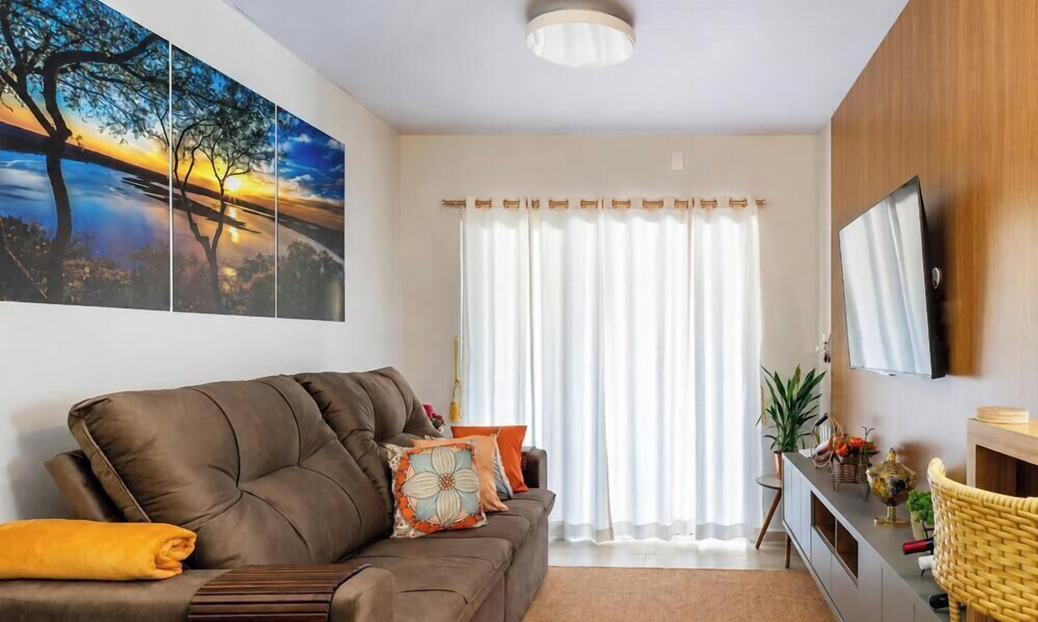 Un salón con un sofá marrón, un televisor de pantalla plana, un cuadro grande y una ventana con cortinas blancas.