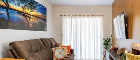Un salón con un sofá marrón, un televisor de pantalla plana, un cuadro grande y una ventana con cortinas blancas.