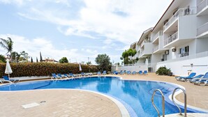 A swimming pool with lounge chairs and umbrellas, surrounded by a white building with balconies.